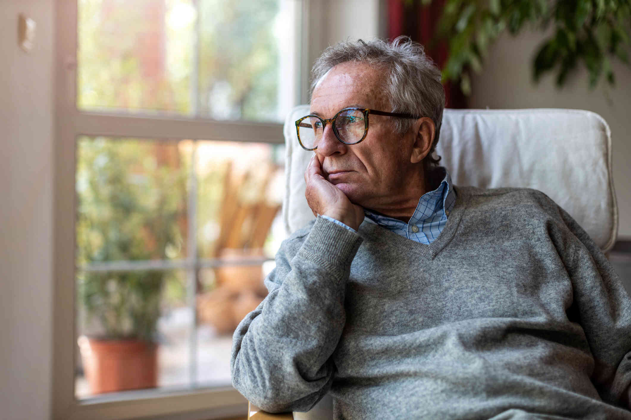 A middle aged man in a grey sweater with glasses sits in an armchair in his home and rests his head in his hand with af ar off expression.